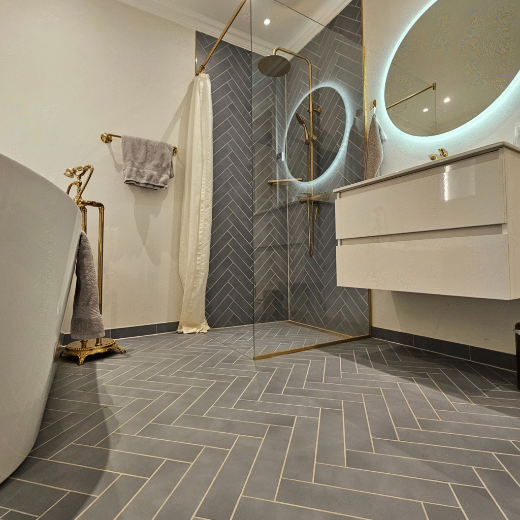 Moderne badeværelse – Brusekabine med grå sildebensfliser, messingarmaturer og rund spejl med lys.