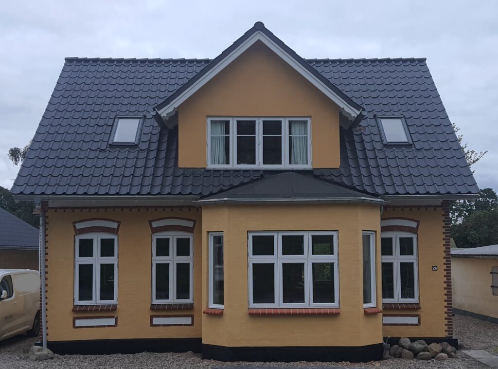 Hus facade efter renovering – Gult murstenshus med nyt sort tegltag og hvide sprossevinduer.
